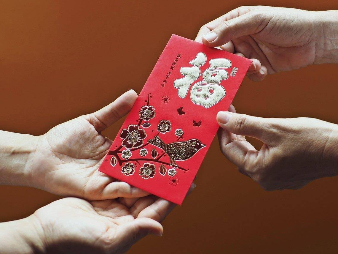 Three hands exchange a red envelope, intricately designed with florals and birds, along with Chinese characters, against a warm orange background—a seamless blend of tradition and meticulous craftsmanship akin to civil engineering outsourcing.